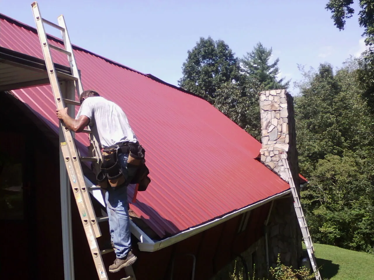 Roofer on a ladder beside a red metal roof and stone chimney — Soffit & Fascia Repair by Roofing Brothers in New Melle